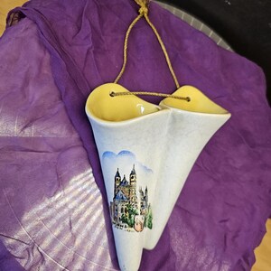 May include: A double-coned ceramic wall vase with a cream-colored exterior and yellow interior. The front features a painted illustration of a cathedral with the text "WORMS" below it. A gold-colored rope is threaded through holes for hanging.