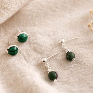 May include: Earrings with green and black malachite stones. The image shows stud and drop earrings, with silver-coloured metal detailing. The malachite stones have a distinctive banded pattern. The earrings are displayed on a neutral fabric.