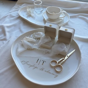 May include: Two white ring boxes with gold accents on a white plate with a gold inscription that reads "I/T" and "Insygnia ilk admiriny". The plate is surrounded by white fabric and pearls.