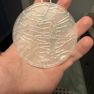 May include: A clear, circular ornament with a textured design. The ornament has a loop at the top for hanging. The design appears to be a landscape or abstract pattern. The ornament is held in a hand.