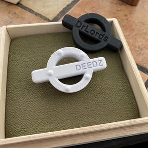 May include: Two white and black round plastic ornaments with the words "Dr. Lords" and "DEEDZ" inscribed on them. The ornaments are in a wooden box with a green felt lining.