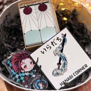 Puede incluir: Un juego de regalo con accesorios temáticos de anime. Incluye un par de pendientes burdeos y blancos, un llavero con un personaje de dibujos animados y un pin con un diseño azul y plateado. Los artículos se presentan en un cuenco decorativo con papel negro.