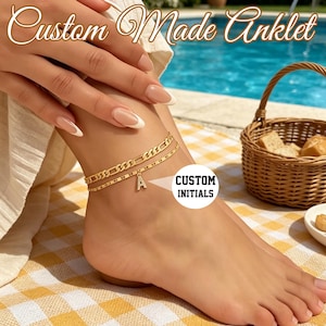May include: A gold-toned double-layered anklet featuring a Figaro chain and a delicate cable chain. The piece includes a single dangling charm in the shape of the letter A. The text Custom Made Anklet and CUSTOM INITIALS appear on the image.