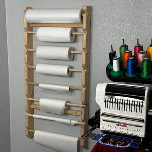 May include: A wooden wall-mounted storage rack holds several rolls of white material. A sewing machine with colorful thread spools is to the right. The machine is white with black accents and the brand name "BERNINA" is visible.