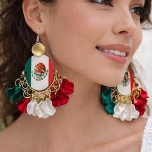 May include: Dangle earrings featuring a circular centerpiece displaying the Mexican flag. The design includes a gold-toned disc at the top, connected to a central oval with the flag, accented by hanging fabric petals in green, white, and red.