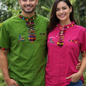 Puede incluir: Dos personas con camisas de manga corta a juego. Una es verde, la otra rosa. Ambas camisas tienen un cuello colorido y un bordado de loro con la palabra "Honduras". Las camisas tienen un escote con cordones.