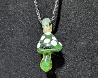 May include: A green glass mushroom pendant with white polka dots hangs from a black cord necklace. The mushroom has a rounded cap and a teardrop-shaped stem.