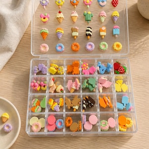 May include: A clear plastic organiser filled with colourful miniature food-themed charms. The charms include ice cream cones, doughnuts, ice lollies, and other sweet treats. The charms are in various colours, including pink, yellow, blue, and orange.