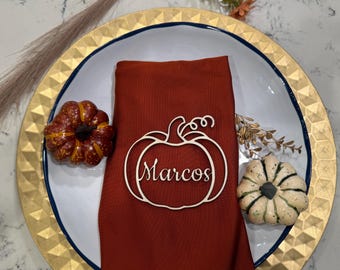 May include: A seasonal table setting featuring a rust-coloured napkin and a pumpkin-shaped place card with the name "Marcos". Decorative pumpkins in orange and white sit on a gold-edged plate.