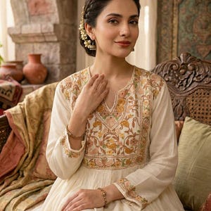 Puede incluir: Vestido crema bordado con escote en V y mangas largas. El vestido presenta intrincados bordados florales en tonos amarillos, naranjas y verdes. La mujer lleva pulseras y pendientes dorados, con el pelo peinado en una trenza.