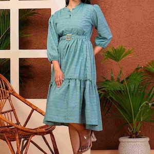 May include: A light teal dress with a subtle grid pattern, featuring three-quarter sleeves and a belted waist with a gold-colored buckle. The dress has a ruffled hem and is paired with clear heeled sandals. The dress is approximately 100 cm in length.