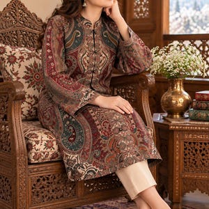 May include: A long-sleeved tunic featuring a complex paisley pattern in shades of deep red, forest green, and cream, paired with solid cream-colored trousers and light-colored, intricately detailed flat shoes.