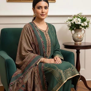 May include: A green ensemble featuring a long-sleeved top and matching pants. The outfit is adorned with gold embroidery and a patterned shawl in shades of brown, green, and red. The woman is seated in a green armchair.