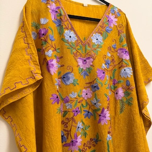May include: Mustard yellow caftan with a V-neck and short sleeves. The caftan features embroidered floral patterns in purple, blue, and green. The sleeve edges are trimmed with a decorative stitch in a contrasting colour.