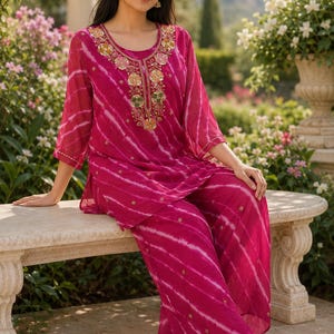 May include: A magenta two-piece outfit featuring a tunic and wide-leg pants with a white diagonal tie-dye pattern. The tunic has three-quarter sleeves and a round neckline adorned with intricate floral embroidery in gold, pink, and green tones.