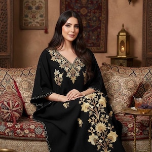 May include: A black caftan dress with gold floral embroidery. The dress has a V-neck and wide sleeves. The embroidery is focused around the neckline and down the front. The dress is worn on a patterned sofa.