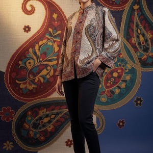 Könnte beinhalten: Eine gemusterte Jacke mit Stehkragen und roten Knopfverschlüssen. Die Jacke hat ein Paisley-Design in Creme, Rot und Türkis. Die Person trägt eine schwarze Hose und Stiefel. Der Hintergrund hat ein großes Paisley-Muster.