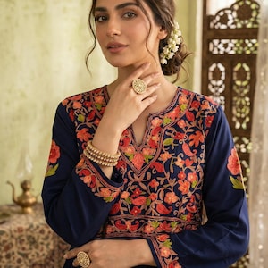 May include: A navy blue dress with long sleeves and detailed floral embroidery in orange, red, and green. The dress has a V-neck and is accessorized with gold rings and bracelets. The style is elegant and detailed, with a focus on craftsmanship.