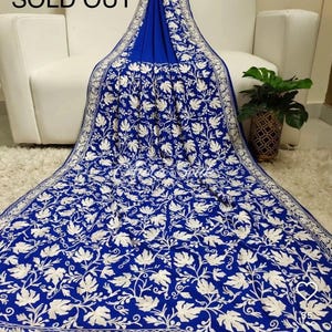 May include: A royal blue textile with intricate white floral embroidery. The fabric is draped over a white sofa and rug, with the words "SOLD OUT" visible in the upper left corner. A potted plant is in the background.
