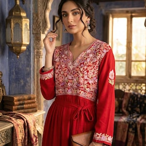 Puede incluir: Vestido rojo con bordado floral en el corpiño y las mangas. El vestido tiene cuello en V y lazo en la cintura. Se sostiene un clutch marrón. El fondo presenta un interior ornamentado con una ventana y una lámpara decorativa.