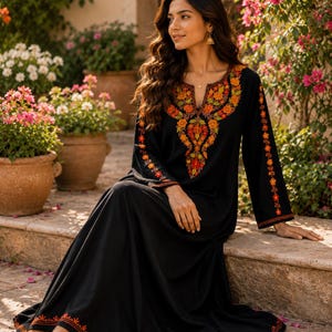 May include: A black floor-length dress featuring a vibrant orange and red floral embroidery pattern around the neckline, down the center of the chest, along the sleeves, and across the bottom hem. The dress has long sleeves and a scoop neckline.