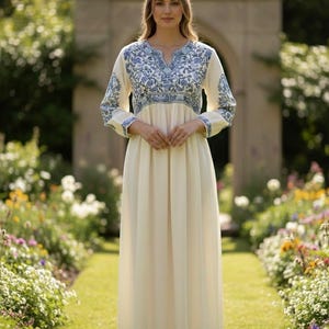 May include: A cream-coloured maxi dress featuring blue floral embroidery on the bodice and sleeves. The dress has a V-neck and long sleeves. The model is standing in a garden with a stone archway in the background.