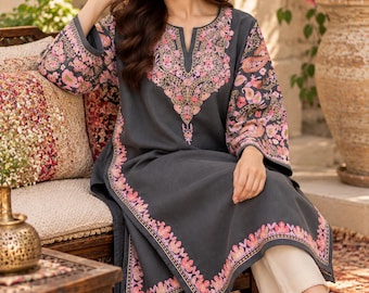 May include: A charcoal gray long-sleeved tunic featuring a notched neckline and intricate floral embroidery in shades of pink, orange, and cream. The embroidery adorns the neckline, sleeve cuffs, and hemline. The tunic is paired with cream-colored trousers.