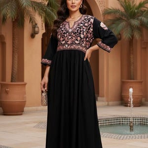 Puede incluir: Vestido largo negro con bordado floral en el corpiño y las mangas. El vestido tiene un escote en V y mangas tres cuartos. Se sostiene un clutch dorado en la mano. El vestido es adecuado para ocasiones especiales.