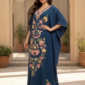 May include: An indigo-coloured caftan dress with a V-neck and short sleeves. The dress features floral embroidery in pink, yellow, and green. The floor-length dress is a flowing design, suitable for warmer weather.