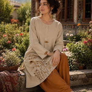 May include: A woman wearing a beige tunic with long sleeves and a brown leather button closure. The tunic features embroidered floral details. She is wearing rust-colored pants and brown leather shoes. The setting is outdoors with flowers and greenery.