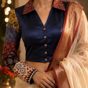 May include: A navy blue button-down blouse with a V-neckline and a red patterned collar. The long sleeves feature a multi-colored floral print in yellow, blue, and black. A cream-colored shawl with a wide copper-toned border is draped over one shoulder.
