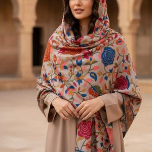 May include: A beige shawl with a floral pattern featuring red, blue, and orange roses and leaves. The shawl is draped over a matching beige dress. The floral design is detailed and colorful, creating a striking contrast against the neutral background.