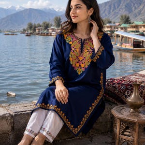 May include: A woman wearing a navy blue embroidered tunic and white pants with floral embroidery. The tunic features a colorful floral design around the neckline and sleeves. The woman is sitting near a body of water.