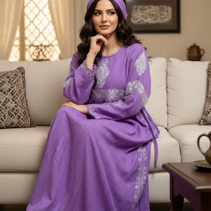 May include: A long, flowing purple dress with intricate silver embroidery on the sleeves and bodice. The dress is paired with a matching head covering. The woman is seated on a cream-coloured sofa in a well-lit room.