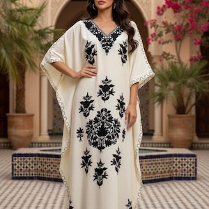 May include: Cream-coloured caftan dress with black floral embroidery. The long, flowing dress features a V-neck and wide sleeves. The embroidery is concentrated around the neckline and down the front. The dress is worn in an outdoor setting.