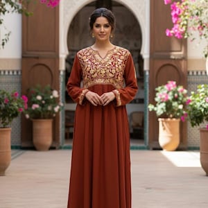 Puede incluir: Un vestido largo color óxido con mangas largas y falda fluida. El corpiño y las mangas están adornados con intrincados bordados dorados. El vestido tiene un escote en V y se usa en un patio con flores rosas.