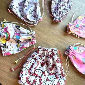 May include: Five drawstring bags with different Hello Kitty designs. The bags are made of white fabric with pink, red, and blue accents. The bags are all tied with white drawstrings.