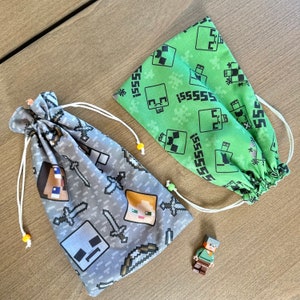 May include: Two drawstring bags with Minecraft character designs. One bag is green with a creeper design and the text "ISSSSSS!" repeated. The other bag is gray with a variety of Minecraft characters, including Steve, Alex, and a creeper.