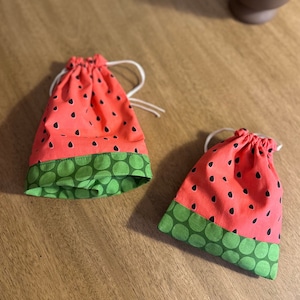 May include: Two small watermelon-patterned drawstring bags with green polka dot trim. The bags are made of fabric and have white drawstrings.