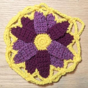May include: A crocheted flower with a purple centre and yellow petals. The flower is surrounded by a yellow crocheted border.