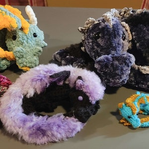 May include: Five crocheted dragon plushies in various colors and styles. The dragons are made with different textures, including fuzzy, smooth, and textured yarn.