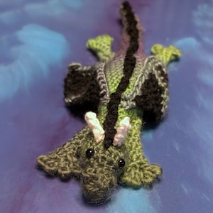 May include: A handmade crochet dragon in shades of green, black, and gray, with a black stripe down its back. The dragon has small black eyes and white and pink horns. The dragon is lying on a blue surface.