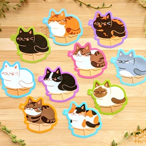 May include: Nine colorful stickers featuring cats sleeping in boxes. Each sticker has a different cat breed and color combination. The stickers are on a wooden background.