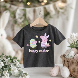 May include: Dark gray t-shirt featuring a cartoon design with a pig and an egg, with the text "hoppy easter". The pig is wearing a dress and holding a basket. The egg has a face and is wearing a hat. The shirt is hanging on a wooden hanger.