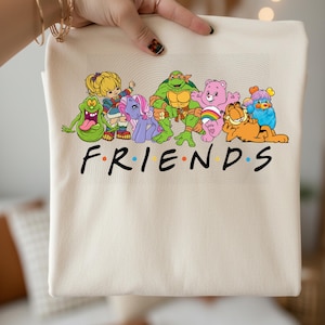 May include: A cream tote bag with a graphic of cartoon characters including a green ghost, a blonde girl, a purple pony, a green turtle, a pink bear, an orange cat, and a blue creature. The word "FRIENDS" is printed in black.