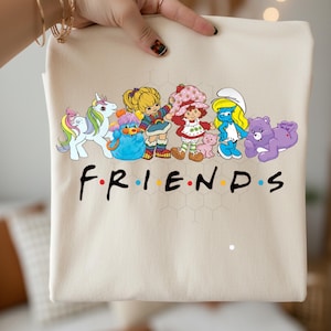 May include: A beige tote bag featuring a colorful graphic of cartoon characters, including a unicorn, Rainbow Brite, Strawberry Shortcake, a Smurf, and a Care Bear. The word "FRIENDS" is printed in black with colorful dots.