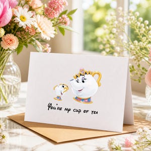 May include: A white greeting card with a cartoon teapot and teacup design, featuring gold detailing. The card reads "You're my cup of tea" in black script. It rests on a brown envelope, with flowers visible in the background.