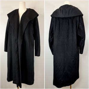 May include: A long, black coat with a large, draped collar. The coat is displayed on a mannequin, showcasing its full length and design. The coat has long sleeves and a flowing silhouette, suitable for outerwear.