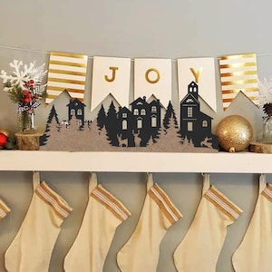 Può includere: Uno scaffale bianco con uno striscione a righe dorate e bianche che recita "JOY" sopra una silhouette nera di una piccola città con alberi e una chiesa. Otto calze beige con righe dorate e bianche pendono sotto lo scaffale.