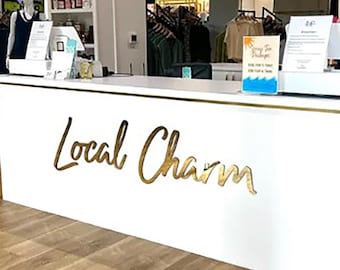 May include: A white retail counter with gold trim and the words "Local Charm" in gold script. The counter is in a store with clothing racks and a mannequin. The floor is light brown wood.